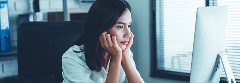 Woman in a white shirt looking at a computer screen, appearing thoughtful.